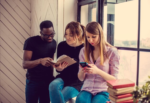 Hombre afroamericano y dos mujeres caucásicas leyendo un libro cerca de la ventana.