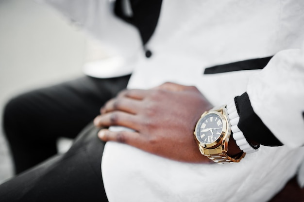 Hombre afroamericano elegante y guapo con traje blanco sentado en un banco Foto de cerca de relojes dorados en la mano
