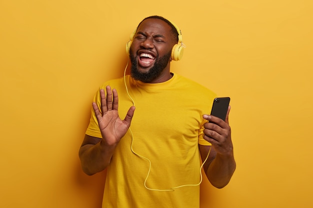 Hombre afroamericano enérgico acerca la palma de la mano a la cámara, usa un teléfono inteligente y auriculares para escuchar la radio o las pistas de audio en la lista de reproducción, mejora el estado de ánimo con su canción favorita