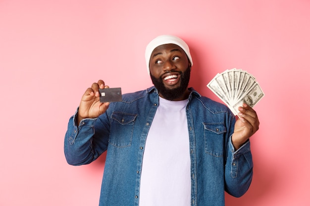 Hombre afroamericano de ensueño mostrando tarjeta de crédito y dólares, pensando en ir de compras y sonriendo, de pie sobre fondo rosa