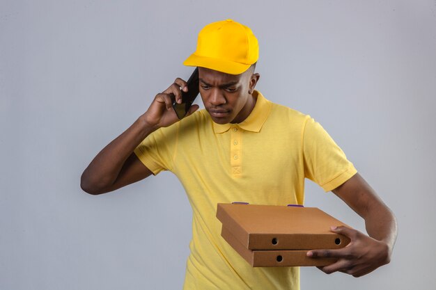 Hombre afroamericano de entrega disgustado en camisa polo amarilla y gorra sosteniendo cajas de pizza mientras habla por teléfono móvil