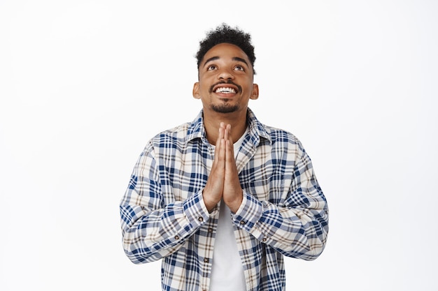 Foto gratuita hombre afroamericano esperanzado rezando a dios, tomados de la mano en un gesto de súplica, mirando hacia el cielo, rogando al señor, de pie con ropa informal contra fondo blanco