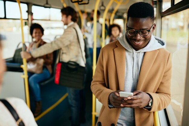 Hombre afroamericano feliz enviando mensajes de texto por teléfono móvil mientras viaja en autobús