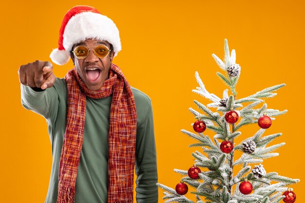 Hombre afroamericano con gorro de Papá Noel y bufanda alrededor del cuello apuntando con el dedo índice a la cámara de pie feliz y emocionado junto a un árbol de Navidad sobre fondo naranja