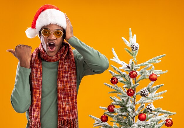 Hombre afroamericano con gorro de Papá Noel y bufanda alrededor del cuello mirando a la cámara asombrado apuntando con el pulgar hacia el lado de pie junto a un árbol de Navidad sobre fondo naranja