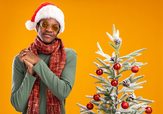 Hombre afroamericano con gorro de Papá Noel y bufanda alrededor del cuello mirando a la cámara sonriendo feliz y positivo tomados de la mano juntos de pie junto a un árbol de Navidad sobre fondo naranja