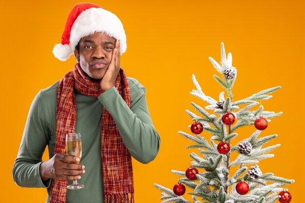 Hombre afroamericano con gorro de Papá Noel y bufanda alrededor del cuello sosteniendo una copa de champán mirando a la cámara cansado y aburrido junto al árbol de Navidad sobre fondo naranja