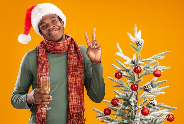 Hombre afroamericano con gorro de Papá Noel y bufanda alrededor del cuello sosteniendo una copa de champán sonriendo alegremente mostrando v-sign junto al árbol de Navidad sobre fondo naranja