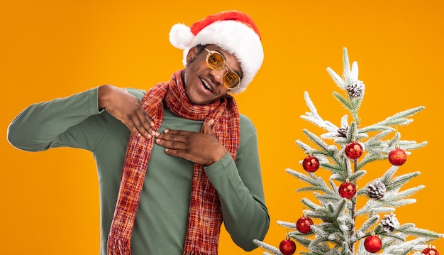 Hombre afroamericano con gorro de papá noel y bufanda alrededor del cuello tomados de la mano en el pecho sentimiento feliz y positivo agradecido de pie junto a un árbol de navidad sobre una pared naranja