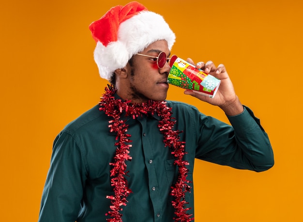 Hombre afroamericano con gorro de Papá Noel con garland con gafas de sol bebiendo de una colorida taza de papel de pie sobre la pared naranja