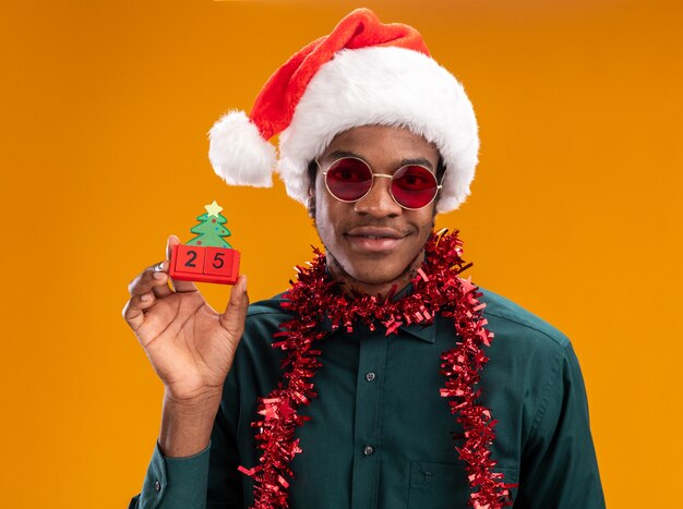 Hombre afroamericano con gorro de Papá Noel con garland con gafas de sol sosteniendo cubos de juguete con fecha veinticinco sonriendo de pie sobre la pared naranja