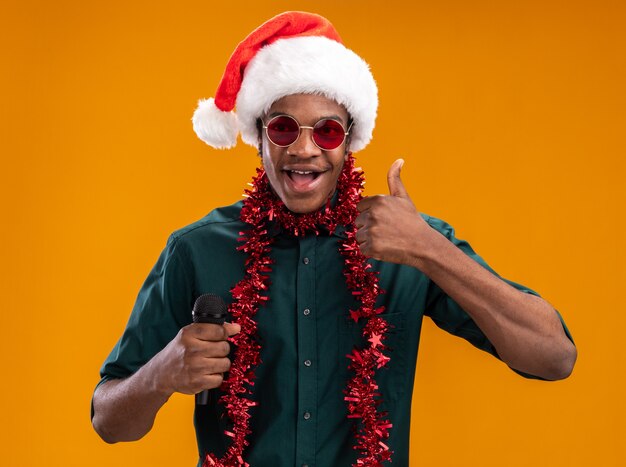 Hombre afroamericano en gorro de Papá Noel con garland con gafas sosteniendo micrófono sonriendo mostrando los pulgares para arriba de pie sobre la pared naranja