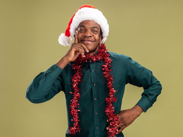 Hombre afroamericano en gorro de Papá Noel con garland mirando a la cámara con una sonrisa en la cara de pie sobre fondo verde