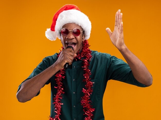 Hombre afroamericano con gorro de Papá Noel con guirnalda con gafas gritando al micrófono con el brazo levantado sobre la pared naranja