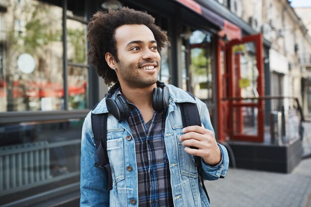 Hombre afroamericano guapo encantador con cerdas y corte de pelo afro mirando a un lado mientras sostiene la mochila y pasea por la ciudad.