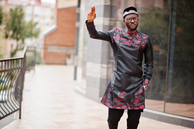 Un hombre afroamericano guapo con gorra de ropa tradicional y anteojos en una ciudad moderna levantó la mano