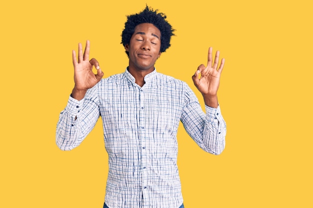 Un hombre afroamericano guapo con el pelo afro vestido con ropa informal se relaja y sonríe con los ojos cerrados haciendo gestos de meditación con los dedos. concepto de yoga