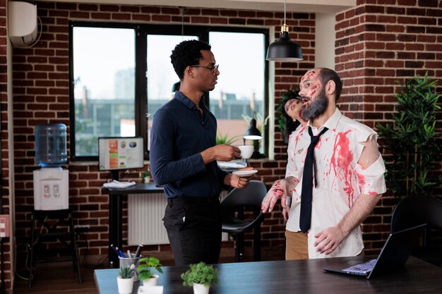 Hombre afroamericano hablando con un zombi en la oficina de negocios, un cadáver espeluznante no muerto charlando con una persona en el lugar de trabajo de inicio. Monstruo macabro de terror malvado con cicatrices sangrientas y decaído.