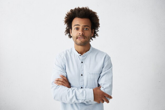 Hombre afroamericano joven asustado en camisa casual manteniendo los brazos cruzados, mordiendo los labios