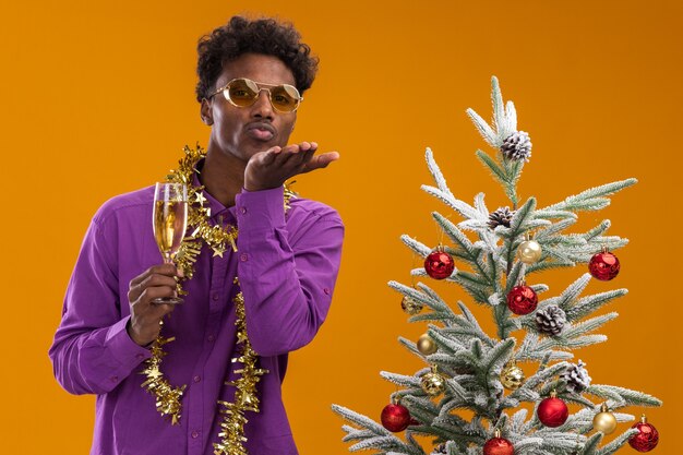 Hombre afroamericano joven confidente con gafas con guirnalda de oropel alrededor del cuello de pie cerca del árbol de Navidad decorado con copa de champán