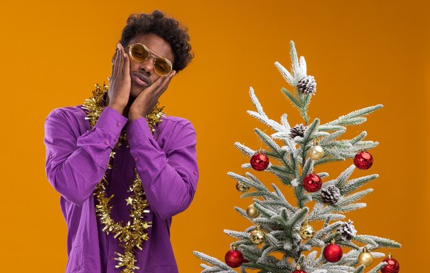 Hombre afroamericano joven confundido con gafas con guirnalda de oropel alrededor del cuello de pie cerca del árbol de Navidad decorado manteniendo las manos en la cara aislada en la pared naranja