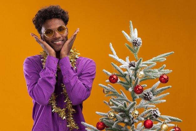 Hombre afroamericano joven emocionado con gafas con guirnalda de oropel alrededor del cuello de pie cerca del árbol de Navidad decorado manteniendo las manos en la cara aislada en la pared naranja