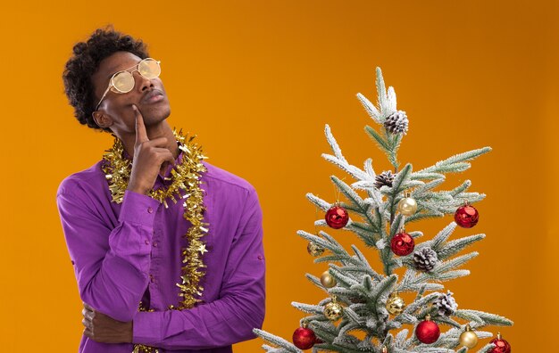 Hombre afroamericano joven pensativo con gafas con guirnalda de oropel alrededor del cuello de pie cerca del árbol de Navidad decorado sobre fondo naranja