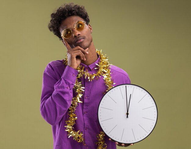 Hombre afroamericano joven pensativo con gafas con guirnalda de oropel alrededor del cuello sosteniendo el reloj manteniendo la mano en la barbilla mirando al lado aislado sobre fondo verde oliva