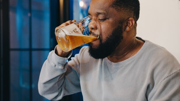 Hombre afroamericano de mediana edad bebiendo cerveza divirtiéndose feliz noche fiesta celebración en línea a través de videollamadas en la sala de estar en casa.