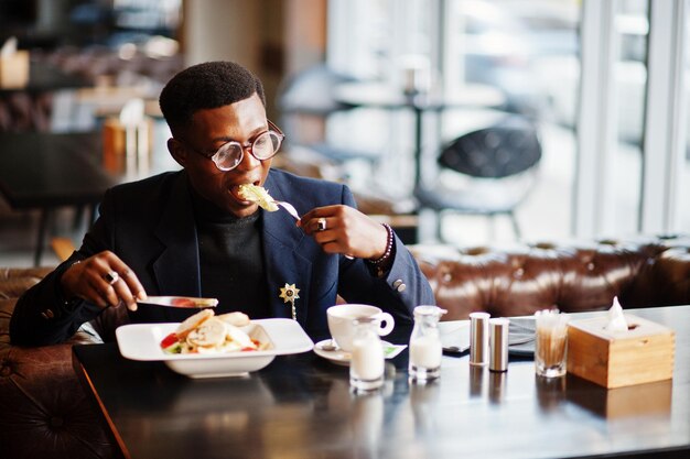 Hombre afroamericano de moda en traje y anteojos sentado en el café y comiendo ensalada