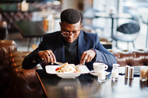 Hombre afroamericano de moda en traje y anteojos sentado en el café y comiendo ensalada