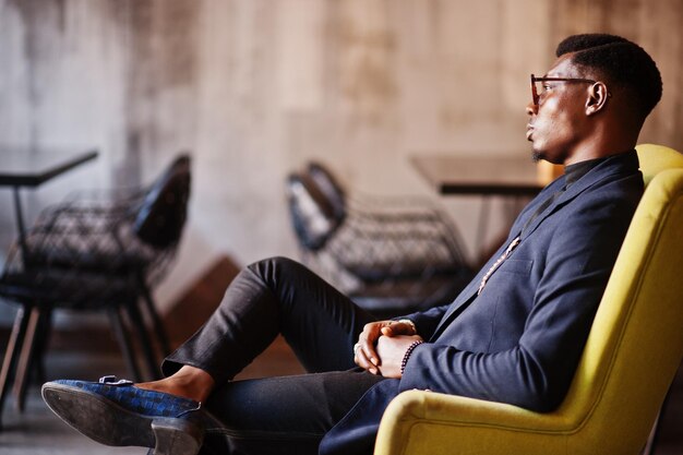 Hombre afroamericano de moda con traje y gafas sentado en una silla en el café