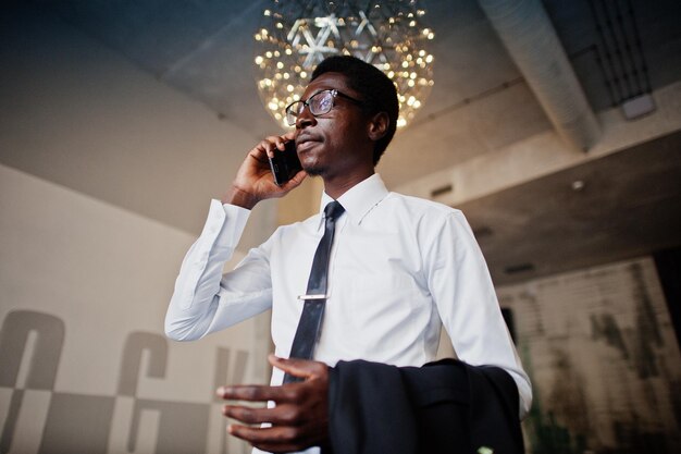 Hombre afroamericano de negocios vestido con camisa blanca, corbata y anteojos en la oficina hablando por teléfono móvil