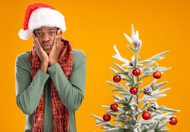 Hombre afroamericano en santa sombrero y bufanda alrededor del cuello mirando a cámara molesto y preocupado de pie junto a un árbol de Navidad sobre fondo naranja