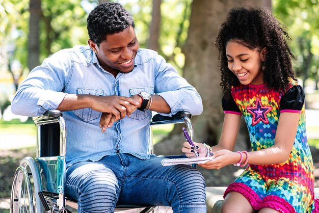 Hombre afroamericano en silla de ruedas disfrutando y divirtiéndose con su hija en el parque.