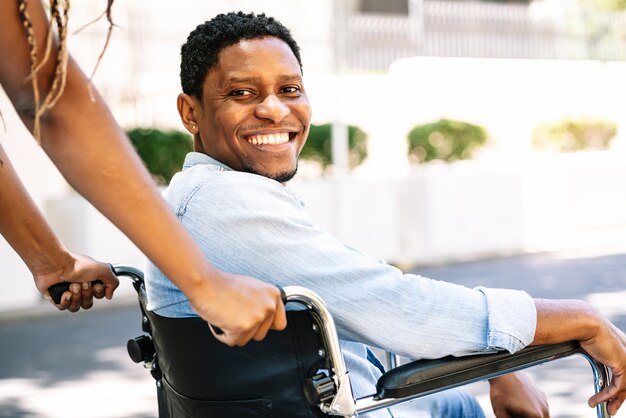 Un hombre afroamericano en silla de ruedas sonriendo y mirando a la cámara mientras su novia lo empuja.
