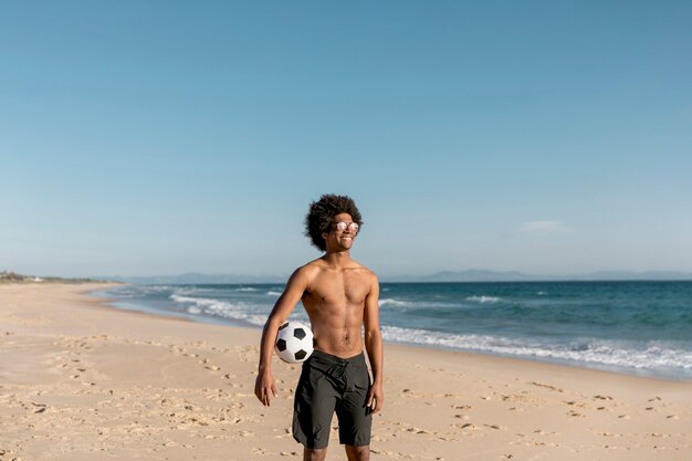 Hombre afroamericano sonriente que se coloca con la bola en la costa