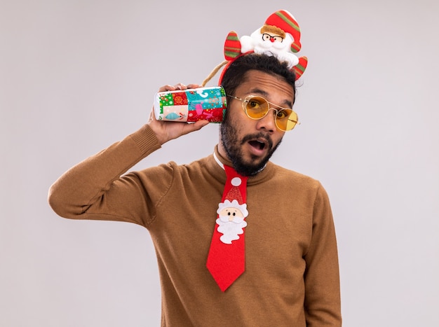 Hombre afroamericano en suéter marrón y santa rim en la cabeza con divertida corbata roja sosteniendo una colorida taza de papel sobre su oreja mirando sorprendido de pie sobre fondo blanco