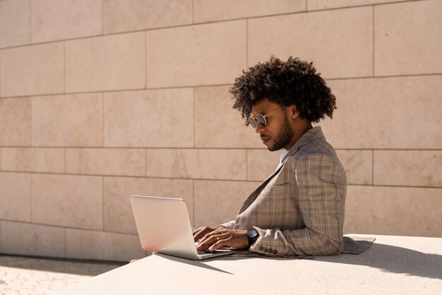 Hombre afroamericano trabajando al aire libre. Hombre con traje y gafas de sol con barba usando laptop. Sentado en la terraza o azoteas. Trabajo, gerente, concepto de tecnología.