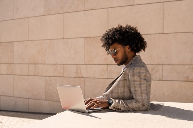 Hombre afroamericano trabajando al aire libre. Hombre con traje y gafas de sol con barba usando laptop. Sentado en la terraza o azoteas. Trabajo, gerente, concepto de tecnología.