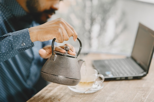 Hombre afroamericano trabajando detrás de una computadora portátil y escribiendo en un cuaderno. Hombre con barba sentado en un café y sirviendo un té.