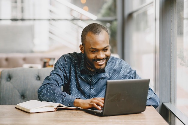 Hombre afroamericano trabajando detrás de una computadora portátil y escribiendo en un cuaderno. Hombre con barba sentado en un café.