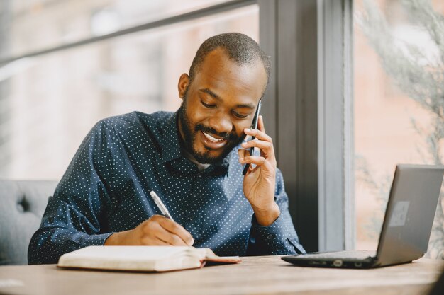 Hombre afroamericano trabajando detrás de una computadora portátil y hablando por teléfono. Hombre con barba sentado en un café.