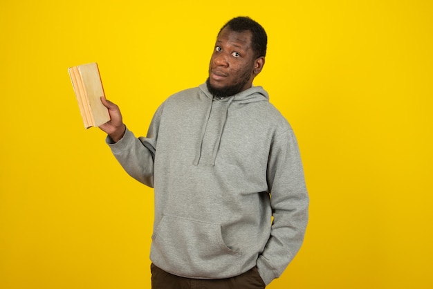 Hombre afroamericano vestido con sudadera gris, con un libro en una mano y la otra en el bolsillo, se encuentra sobre una pared amarilla.