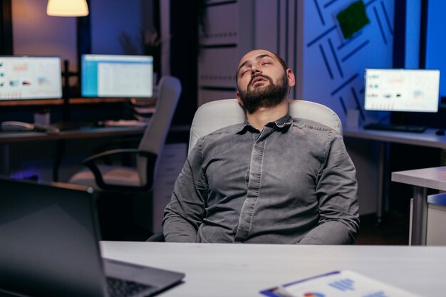 Hombre agotado con exceso de trabajo duerme en una silla en la oficina vacía. Empleado adicto al trabajo que se queda dormido porque trabaja tarde en la noche solo en la oficina para un proyecto importante de la empresa.