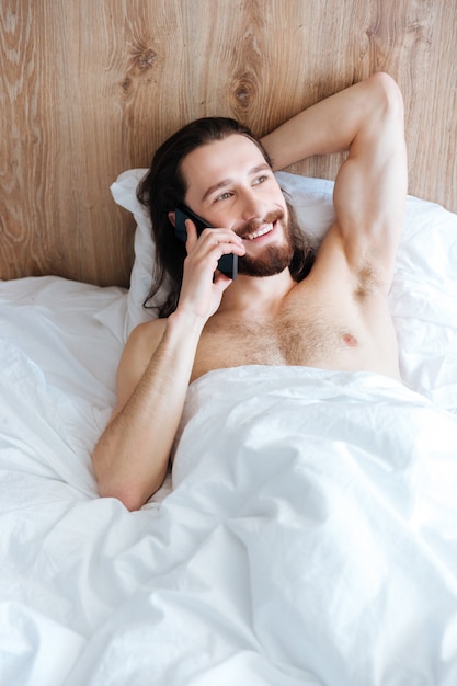 Hombre alegre acostado en la cama y hablando por teléfono celular