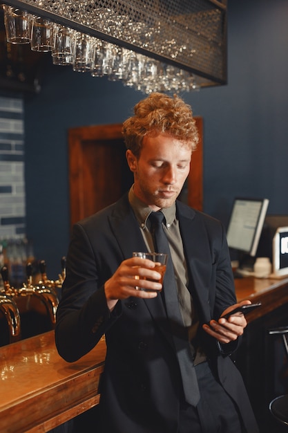 Hombre alegre confiado sentado en la barra del bar, bebiendo cerveza y redes en el teléfono inteligente.