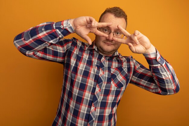 Hombre alegre con gafas y camisa a cuadros sonriendo alegremente mostrando el signo v cerca de los ojos de pie sobre la pared naranja