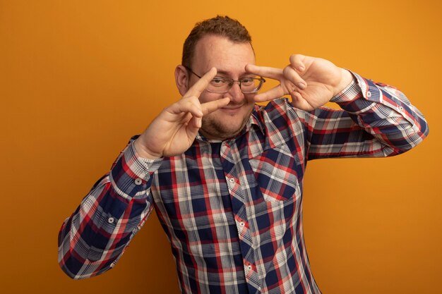 Hombre alegre con gafas y camisa a cuadros sonriendo alegremente mostrando el signo v cerca de los ojos de pie sobre la pared naranja