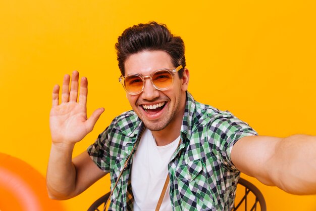 Hombre alegre con gafas naranjas toma selfie y muestra su palma en un espacio aislado.
